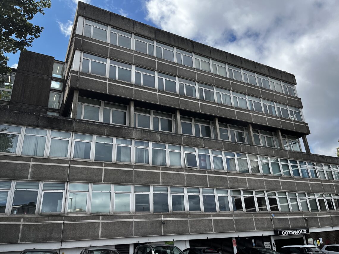A large, grey, concrete office building with four storeys of windows is shown under a partly cloudy sky. Cars are parked in front at ground level, where a sign reading Cotswold is visible above a parking area.