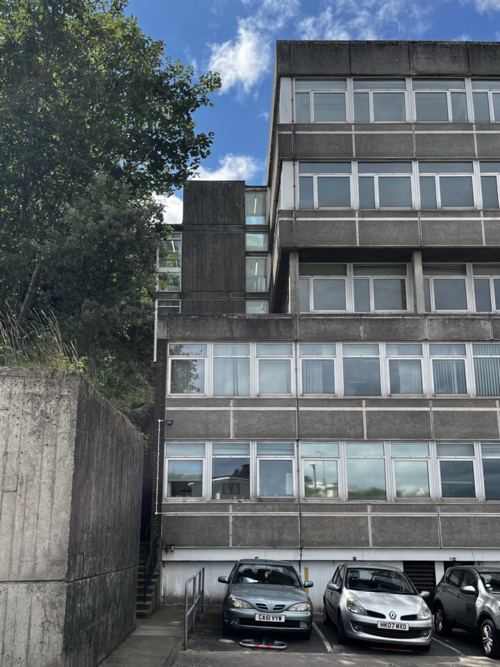 A grey concrete office building with rows of windows is shown. Three cars are parked in front, and a stairway is seen at the left. Trees and blue sky with clouds appear above the roof, and sunlight highlights parts of the building.