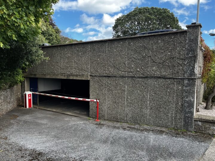 A low, flat-roofed concrete car park with a red and white barrier at the entrance. Trees and bushes partially surround the building, and the sky above is partly cloudy with patches of blue visible. Gravel covers the foreground ground.