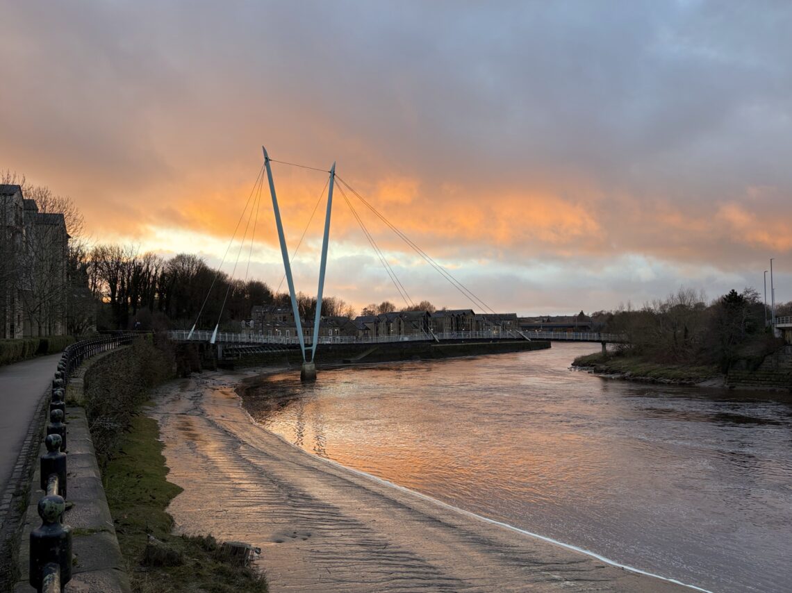 A modern cable-stayed footbridge stretches over a calm river at sunset, with orange and pink clouds filling the sky. Trees and buildings line the riverbanks, and a paved walkway follows the water’s edge on the left side of the image.