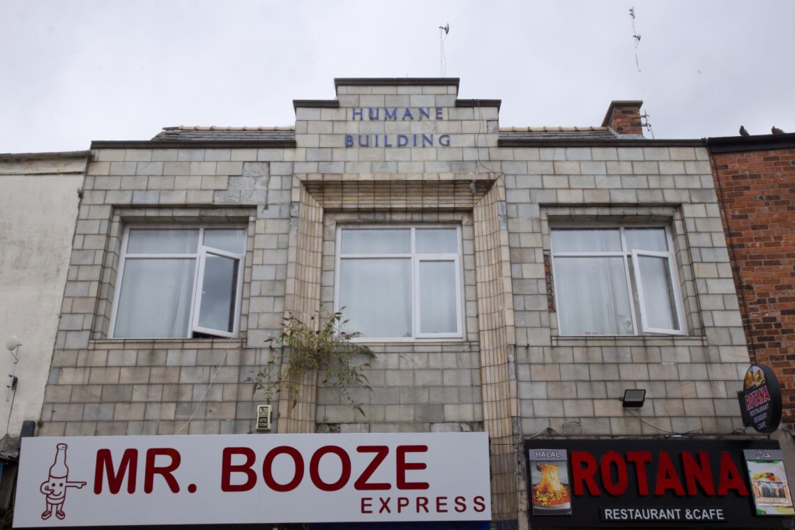 A grey brick building with “HUMANE BUILDING” inscribed above two large windows. Below, a “MR. BOOZE EXPRESS” off-licence sign with a bottle logo and “ROTANA RESTAURANT & CAFE” sign are visible. Some plants grow near the windows.
