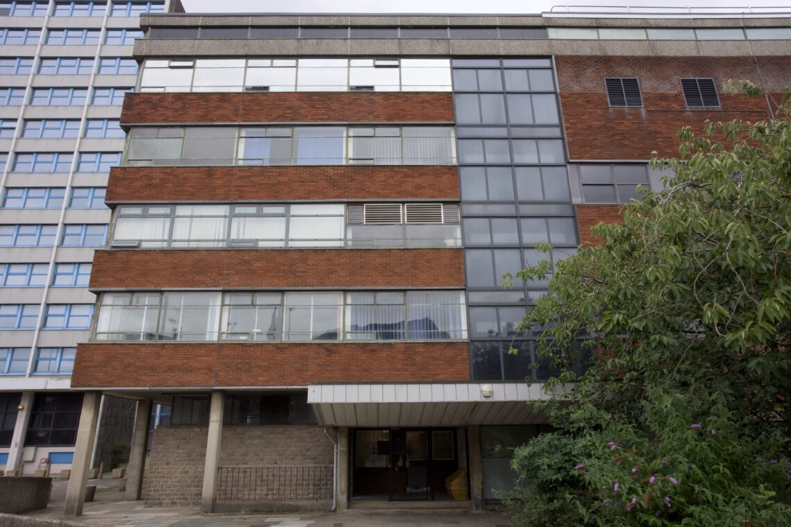 A mid-century modern building with red brick and large glass windows stands next to an older blue-panelled structure. The building has four storeys, a covered entrance, and greenery growing in front on the right side.