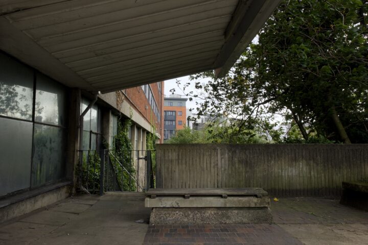 A concrete shelter with a slanted roof overlooks a weathered bench and brick pavement. Overgrown plants climb nearby fences. In the background, red and grey block of flats and dense green trees are visible under a cloudy sky.
