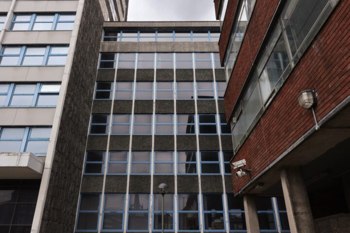 A view upward between two buildings shows a dark, mid-century office block with blue-framed windows. The sky is overcast. The building on the right is red brick, while the left building has a white exterior. A CCTV camera and a lamp are visible.