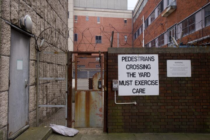 A gated alleyway with brick and concrete walls, topped with barbed wire, features a large white sign reading PEDESTRIANS CROSSING THE YARD MUST EXERCISE CARE. An old metal door and a small warning sign are also visible. A pillow lies on the ground.