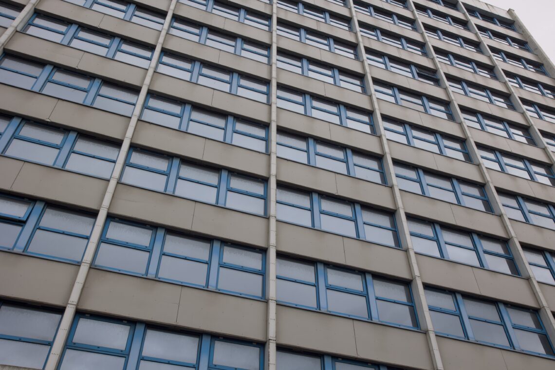 A close-up upward view of a tall building facade featuring multiple rows of rectangular blue-framed windows set in beige panels, with vertical structural beams between the window columns. The perspective creates a slightly distorted, diagonal appearance.