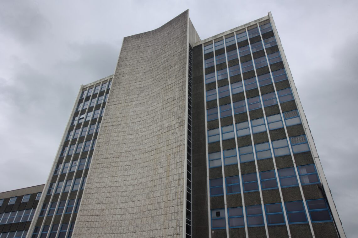 A tall, modern building with a central curved stone façade flanked by two rectangular wings featuring large grid-like windows. The sky is overcast with grey clouds, enhancing the stark architectural lines and highlighting the building’s geometric design.