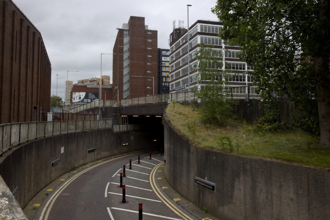 A curved urban road leads downhill into a tunnel beneath concrete and brick buildings. Tall offices and flats rise above, with greenery on the right embankment. Yellow road markings and black bollards guide traffic under a cloudy sky.