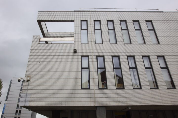 A modern, rectangular white building with vertical narrow windows in staggered rows. The facade is covered with rectangular tiles, and part of the upper corner is open. The sky is overcast and grey.