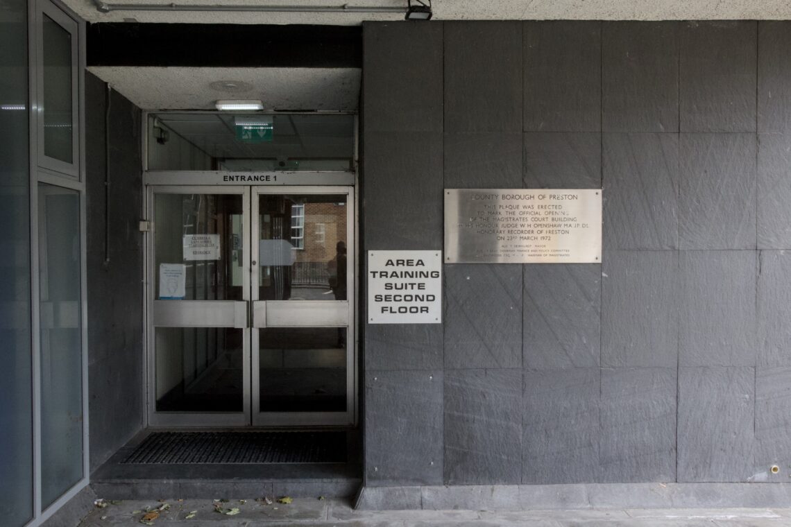 Entrance to a building with double glass doors, a sign reading Area Training Suite First Floor, and a metal plaque on the wall. The plaque lists the County Borough of Preston and council information. The area is shaded and concrete.