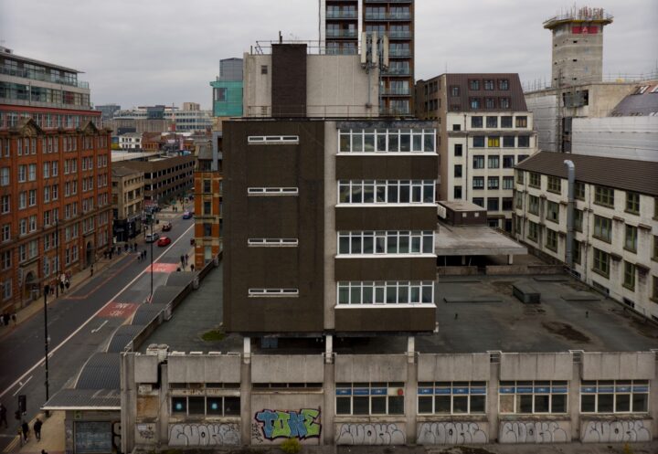 A cityscape view shows a blocky, modernist grey building with many windows. Surrounding it are older brick and contemporary buildings. The nearest wall at street level features colourful graffiti. The scene is cloudy with sparse traffic on the roads.