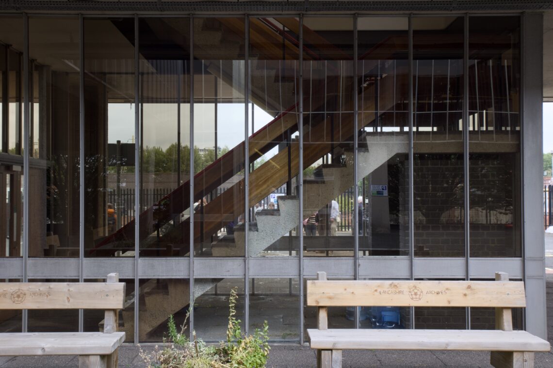 A glass wall reveals a concrete staircase with a wooden handrail inside a building. Two wooden benches are in the foreground outside, with some plants between them. Reflections and vertical window dividers are visible on the glass.