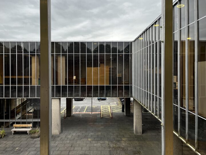 View through a window of a modern building with large glass panels and a rectangular courtyard. The courtyard has paving, parking spaces with yellow lines, pillars, a bench, and trees reflected in the reflective glass façade. Sky is overcast.