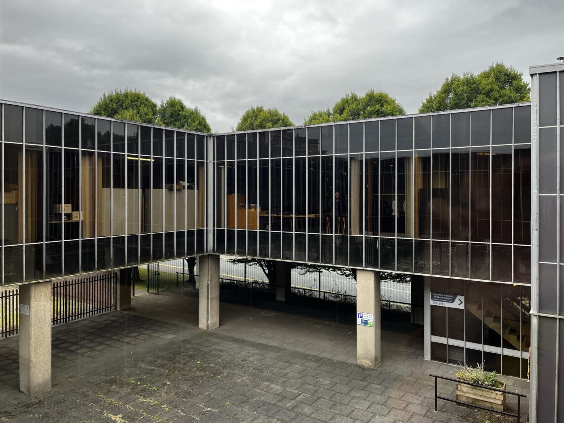 A modern building with large, reflective glass windows forms an elevated walkway supported by pillars. Below, a paved courtyard with a bench and planter is visible. Lush green trees and a cloudy sky are seen in the background.