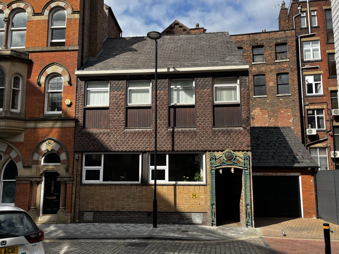 A two-storey brick building with tall windows, wooden and shingle details, and a decorative green metal archway beside a black garage door. The building is flanked by older red-brick structures and a black streetlamp stands in front. Parked cars line the street.