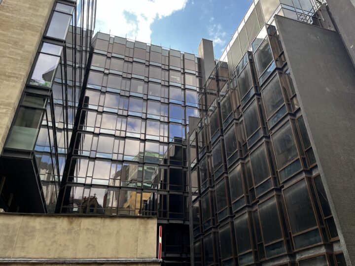 Modern office building with large glass windows reflecting the sky and surrounding structures. The façade features black metal frames and beige concrete sections. The sky above is partly cloudy, and the perspective looks upward from a street level angle.