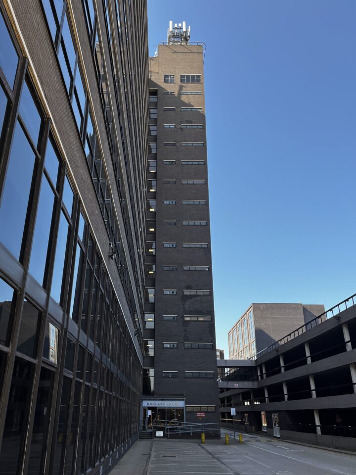 A tall, dark brick office building with many narrow windows stands in bright sunlight against a clear blue sky. Reflections appear in the glass windows on the left. A car park is attached at right, and the area in front is mostly empty pavement.