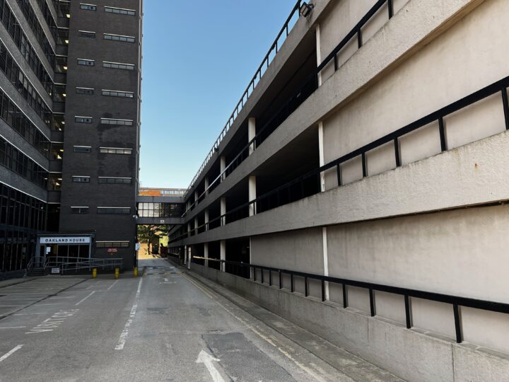A narrow, empty street runs between a dark, tall office block on the left and a beige multi-storey car park on the right. The sky is clear and blue, and a pedestrian bridge connects the two buildings above the street in the background.