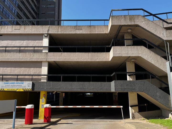 A multi-storey concrete car park with three visible floors and an exterior staircase on the right. A striped barrier blocks vehicle entry, and two red-and-white bollards are at the entrance. Sunlight casts shadows; a sign reads Oakland House.