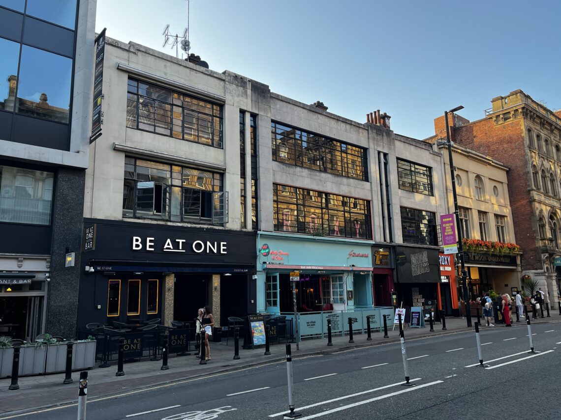 A city street with modern and older buildings. Visible venues include BE AT ONE bar with black signage, a bright blue restaurant with pink and red details, and other shops. People walk on the pavement. The sky is clear and the road has bollards.
