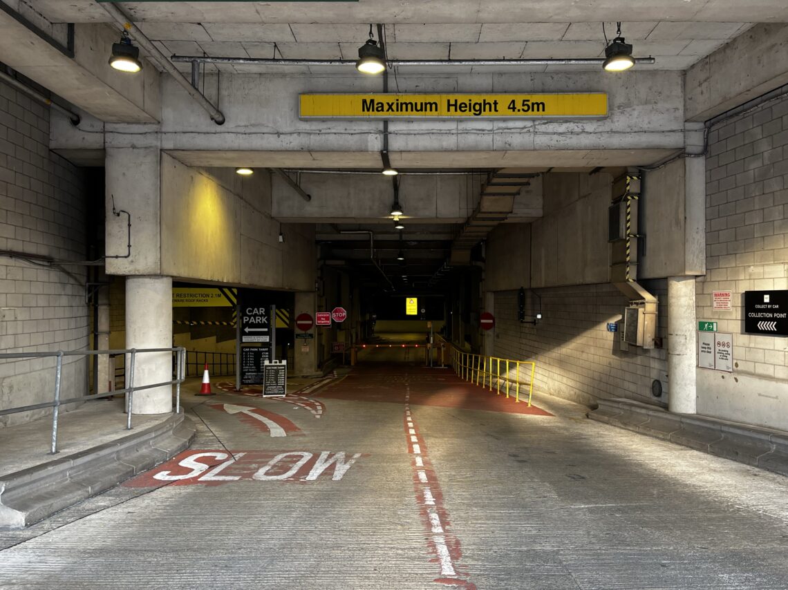 A concrete car park entrance with overhead “Maximum Height 4.5m” sign, SLOW painted on the ramp, red and white bollards, stop signs, and arrows directing cars. Yellow safety railings and various signs are visible along the grey walls.