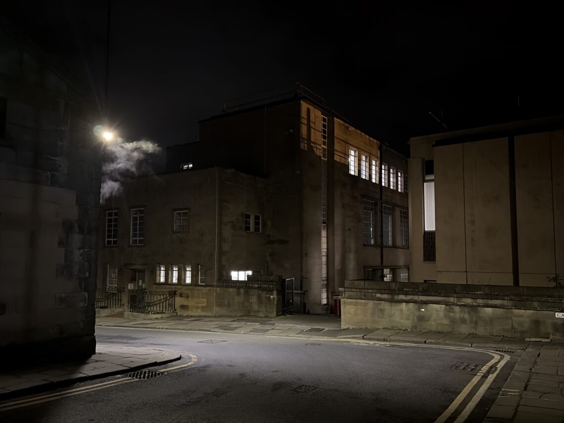 A dimly lit street corner at night with old stone buildings. Warm light glows from upper windows, while a streetlamp casts a yellow light. Steam or smoke drifts near one building. The street is empty and shadows cover much of the scene.
