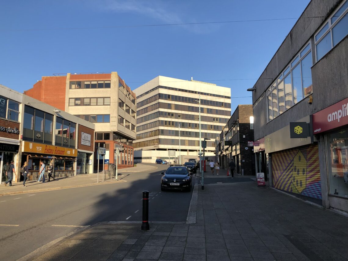 A quiet urban street flanked by mid-rise office buildings and shops, including a sushi restaurant and an amplifier shop with a colourful shutter. A few people walk on the pavement. One car is parked on the street under a clear blue sky.