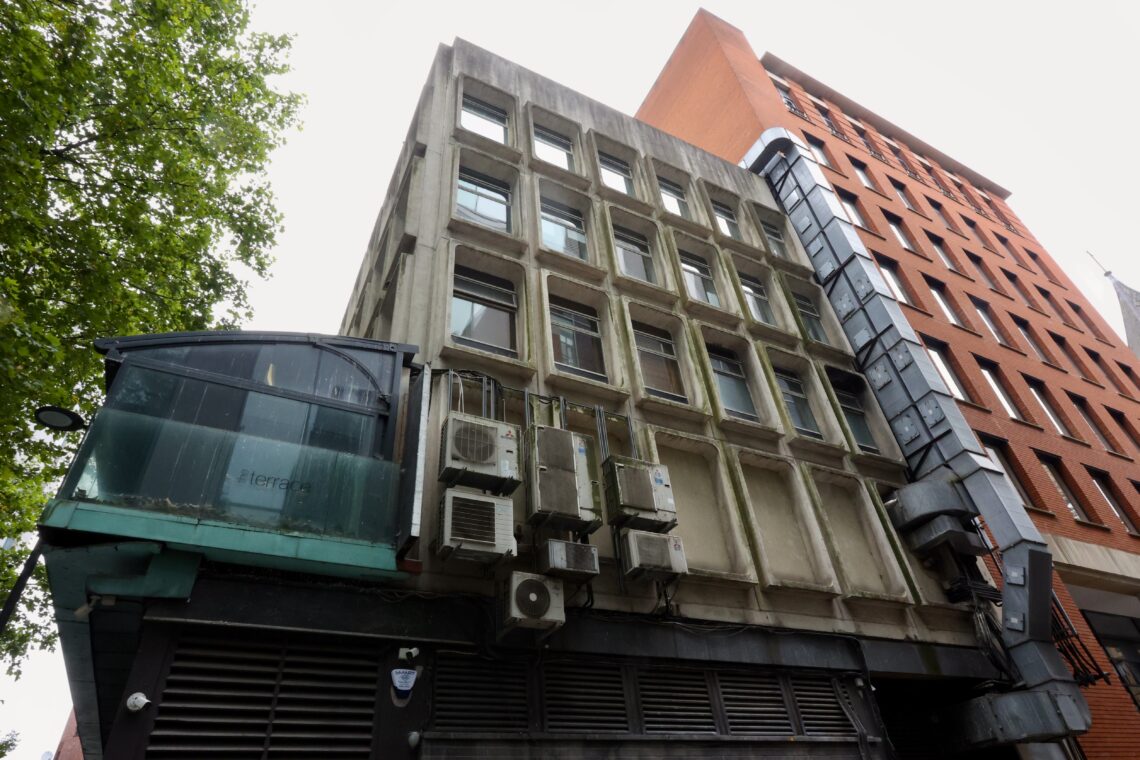A low-angle view of two connected buildings: one grey with square windows and many air conditioning units attached, the other taller, red-brick with a glass and metal staircase running along its side. A green tree is visible on the left edge against a cloudy sky.