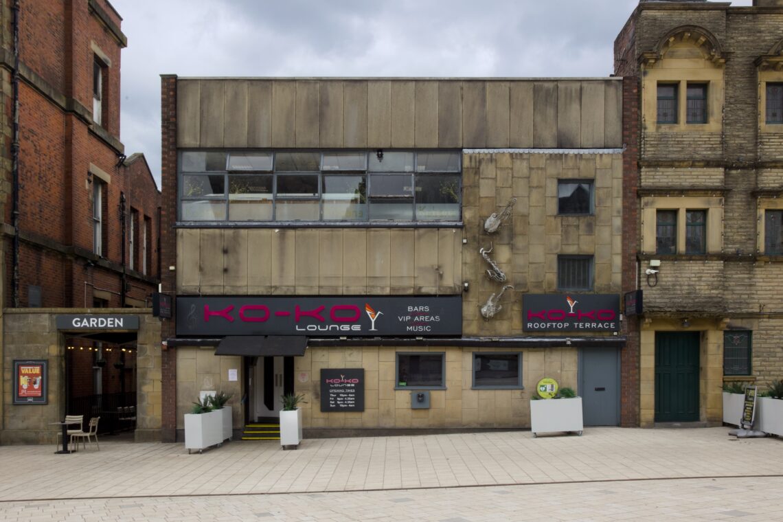 A beige, two-storey building houses KO-KO Lounge bar. It has square windows, modern black signs with red and white text, and a silver martini glass logo. The entrance is flanked by plants and posters. To the left is a garden area; the pavement is pale stone.