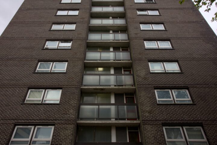 A low-angle view of a brown brick block of flats, showing several storeys with rows of rectangular windows and balconies in the centre. The sky above is cloudy and the branches of a tree are visible in the top right corner.