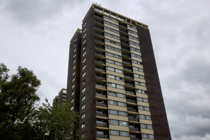 A tall, dark-brown concrete block of flats with many balconies and windows stands against a cloudy, overcast sky. Green trees are visible at the base in the foreground, and the building appears slightly weathered.