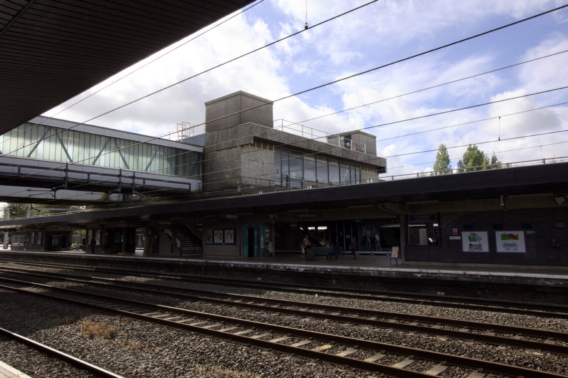 A modern railway station with multiple railway tracks in the foreground and a platform with a large concrete building above. There is a footbridge with glass panels, people waiting on the platform, and posters on the walls. Trees and a partly cloudy sky are visible.
