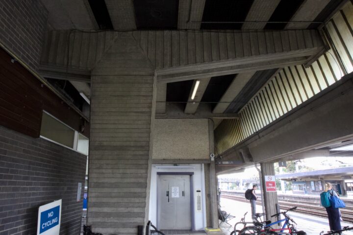 A concrete railway station platform with a lift at the centre. Bicycles are parked nearby, and a cyclist with a blue jacket stands beside one. Signs include “NO CYCLING.” Railway tracks and another platform are visible through the yellow-tinted windows.