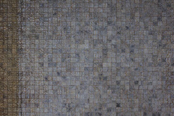 A close-up view of a wall covered with small square greyish-blue tiles. The tiles have a slightly rough, textured surface, and some areas on the left show discolouration or staining in a brownish tone. The overall pattern appears grid-like and repetitive.