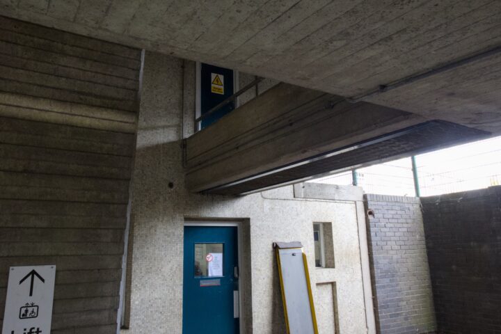 A blue door set into a concrete wall is partially blocked by an overhead ramp. A yellow warning sign and a no smoking sign are visible on the door. A propped-up yellow platform and a sign with an upward arrow and text are also seen nearby.