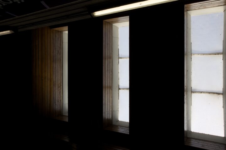 Three tall, narrow windows with frosted glass let in diffused light, surrounded by wooden panelling. The surrounding room is dark, highlighting the bright, vertical rectangles of the illuminated window panes.