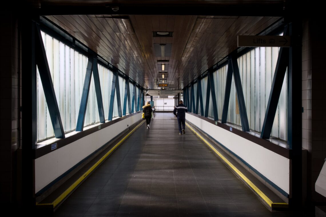 Two people walk away down a long, enclosed pedestrian walkway with frosted windows and blue steel beams. The corridor is brightly lit, with a dark floor, yellow safety lines, and a sign above pointing to “Lifts”.