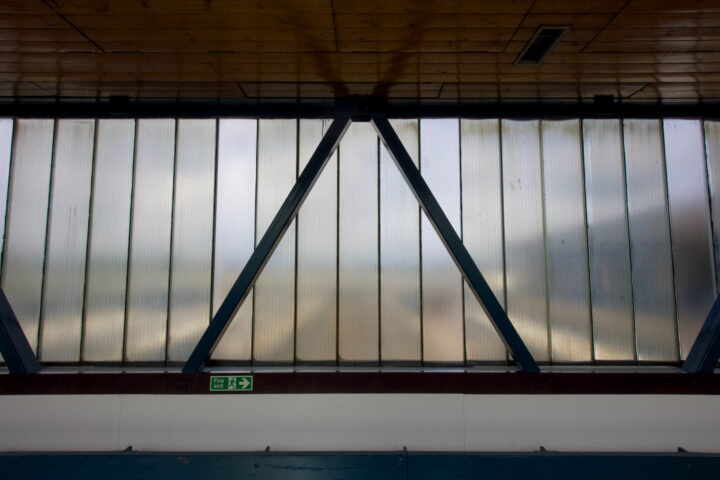 Frosted glass wall with vertical panels and blue triangular metal supports. A small green emergency exit sign with a running figure and arrow is mounted on the white wall below. The wooden ceiling is partially visible at the top.
