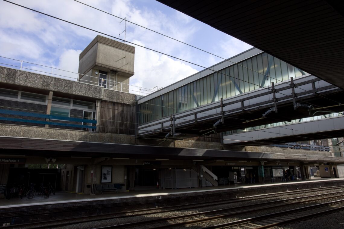 A modern railway station with concrete and glass architecture, featuring a glass-enclosed pedestrian bridge above railway tracks, platforms below with people and bicycles, and a square tower structure in the background under a partly cloudy sky.