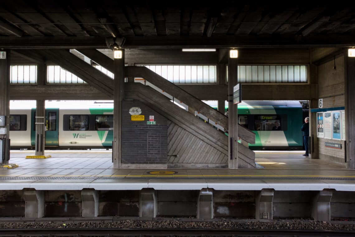 A modern railway station platform with a concrete staircase in the centre, a green and white train stopped in the background, and a person standing near the train doors. Overhead lights and signage are visible under the wooden roof.