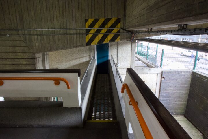 A narrow metal walkway with a blue door at the end, bordered by yellow and black hazard stripes above. The walkway is flanked by concrete and railings with orange handrails, located inside a car park with some daylight visible outside.