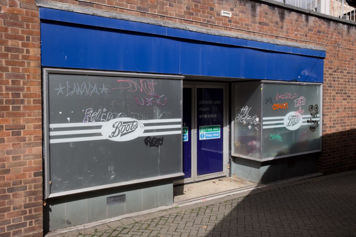 A closed Boots shop with a blue awning and frosted windows covered in graffiti. The Boots logo is visible on both windows. A sign on the door says “Please use main entrance.” The building exterior is brick and the ground is paved.