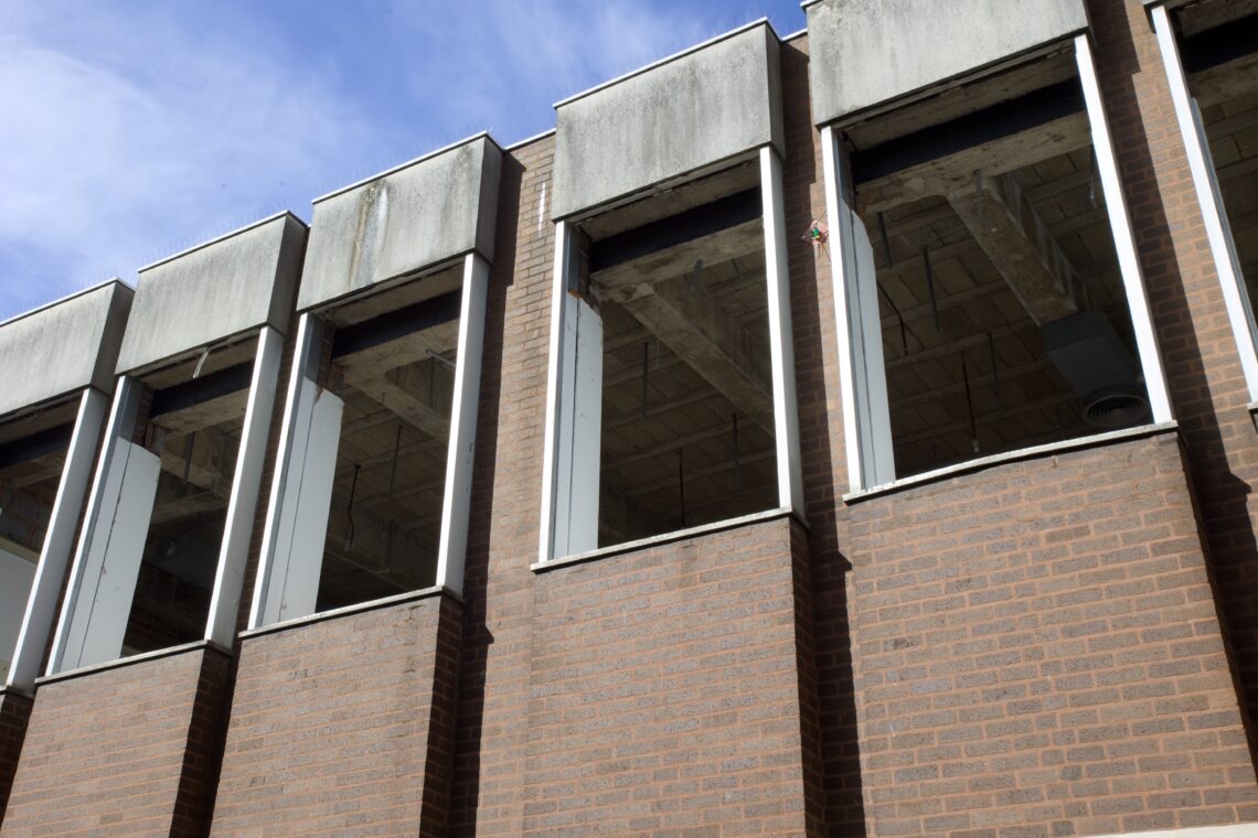 The image shows a brick building with tall, narrow, rectangular windows that have no glass, revealing an unfinished concrete ceiling inside. The upper sections of the building show discolouration and grime. The sky above is clear and blue.