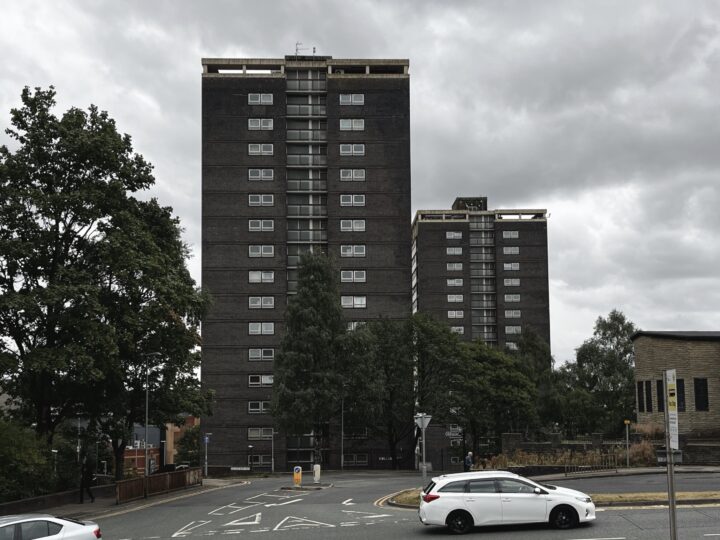 A cloudy day showing two tall, dark high-rise block of flats with many windows. A white car is driving along a curved street in the foreground, with trees and a few low buildings around the towers. The sky is grey and overcast.