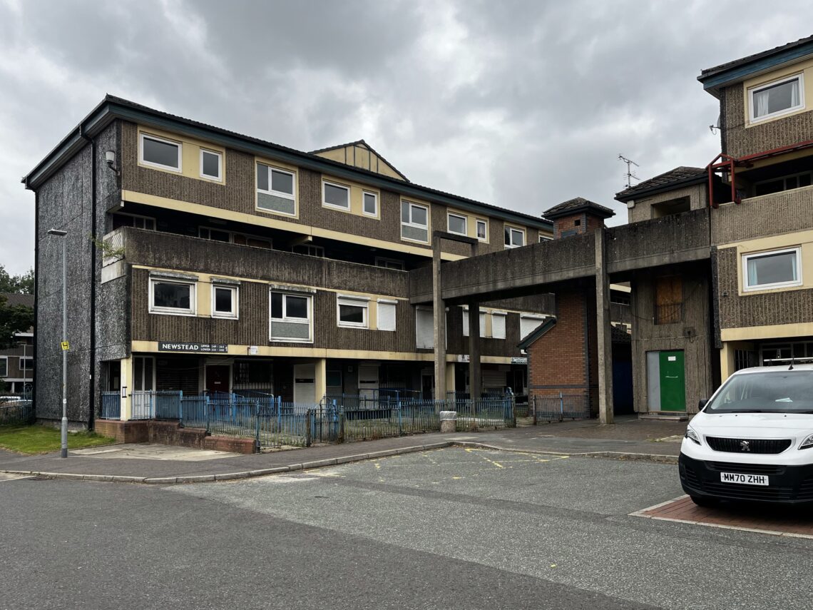 A three-storey, concrete block of flats with weathered walls and boarded-up windows, featuring a raised walkway connecting to another building. A white car is parked in front on a grey, overcast day. Blue railings and empty pavement are visible.