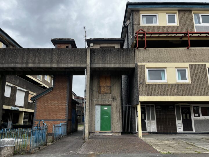 A run-down block of flats with boarded-up windows and doors, stained concrete, a green door, and an elevated walkway connecting two sections. The sky is overcast, and the area looks neglected with no people present.