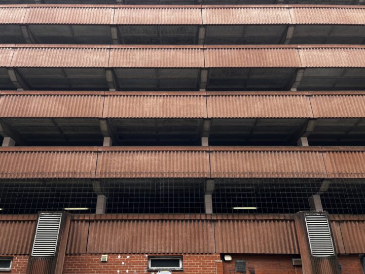 A multi-storey car park with brown, corrugated metal panels and concrete floors. The lower level features brickwork, metal vents, and small windows. Vertical and horizontal lines create a grid-like, repetitive architectural pattern. No cars are visible.