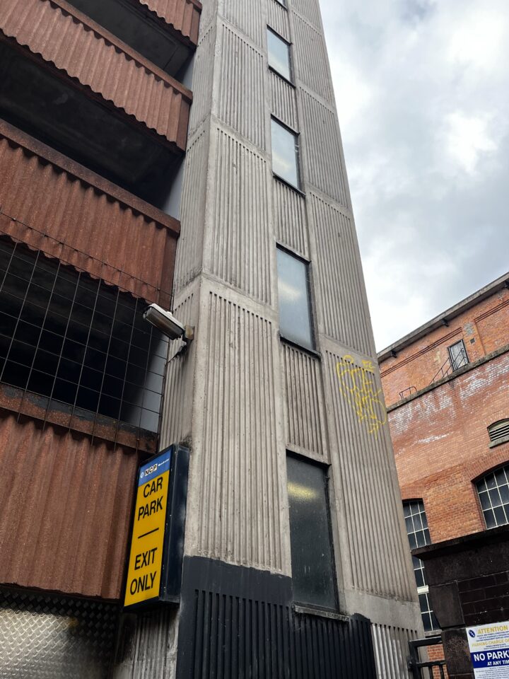 A tall, narrow concrete stairwell with vertical grooves and tall windows stands next to a multi-storey car park. A yellow “Car Park Exit Only” sign is mounted on the wall. The sky is overcast and part of a red brick building is visible.