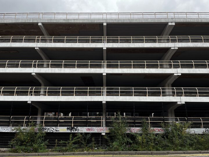 A multi-storey concrete car park with exposed floors and railings. The lower level is covered in colourful graffiti. Overgrown plants line the base, and the sky above is cloudy. The structure appears empty and neglected.