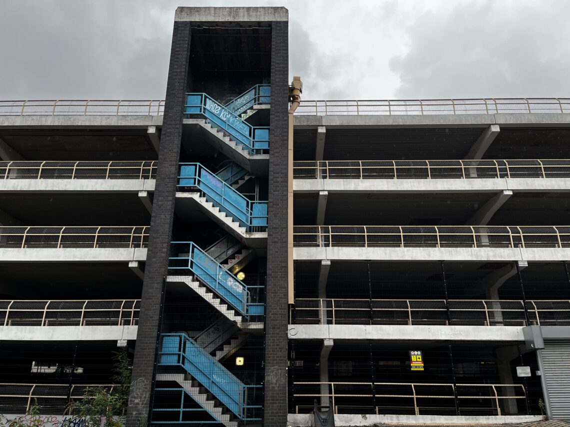A multi-storey car park with exposed concrete, black columns, and bright blue stair railings. The central staircase is visible, zigzagging upward. Overhead, the sky is cloudy and grey. A tall, rusted lamp post stands in front of the building.